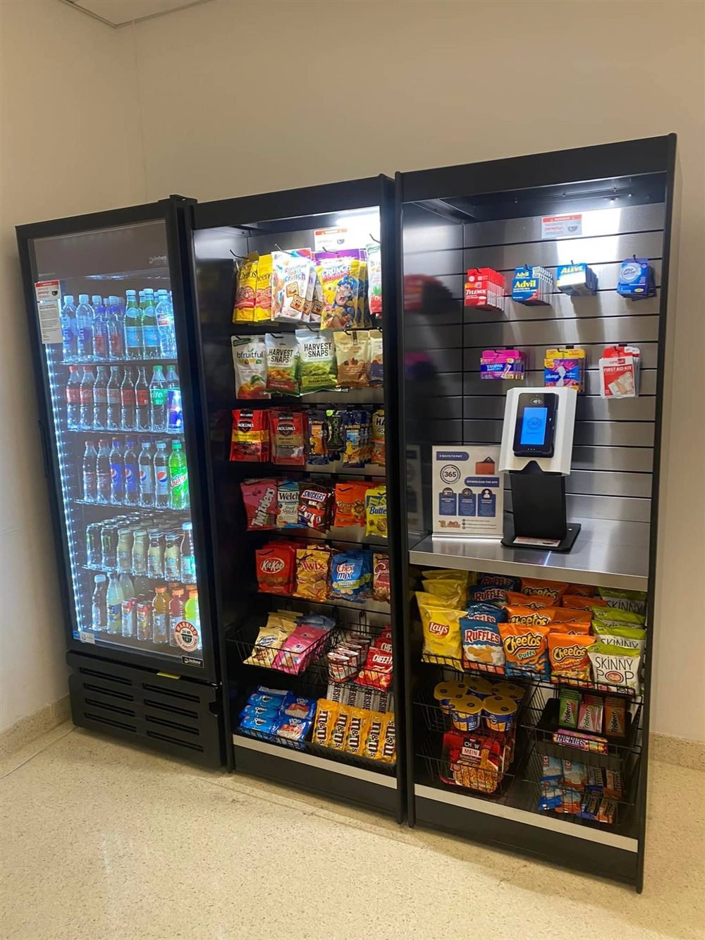 Cashless self-checkout kiosk beside a glass-door cooler and snack shelves